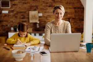 Happy mother working on a computer while her daughter is writing in notebook and learning at home.